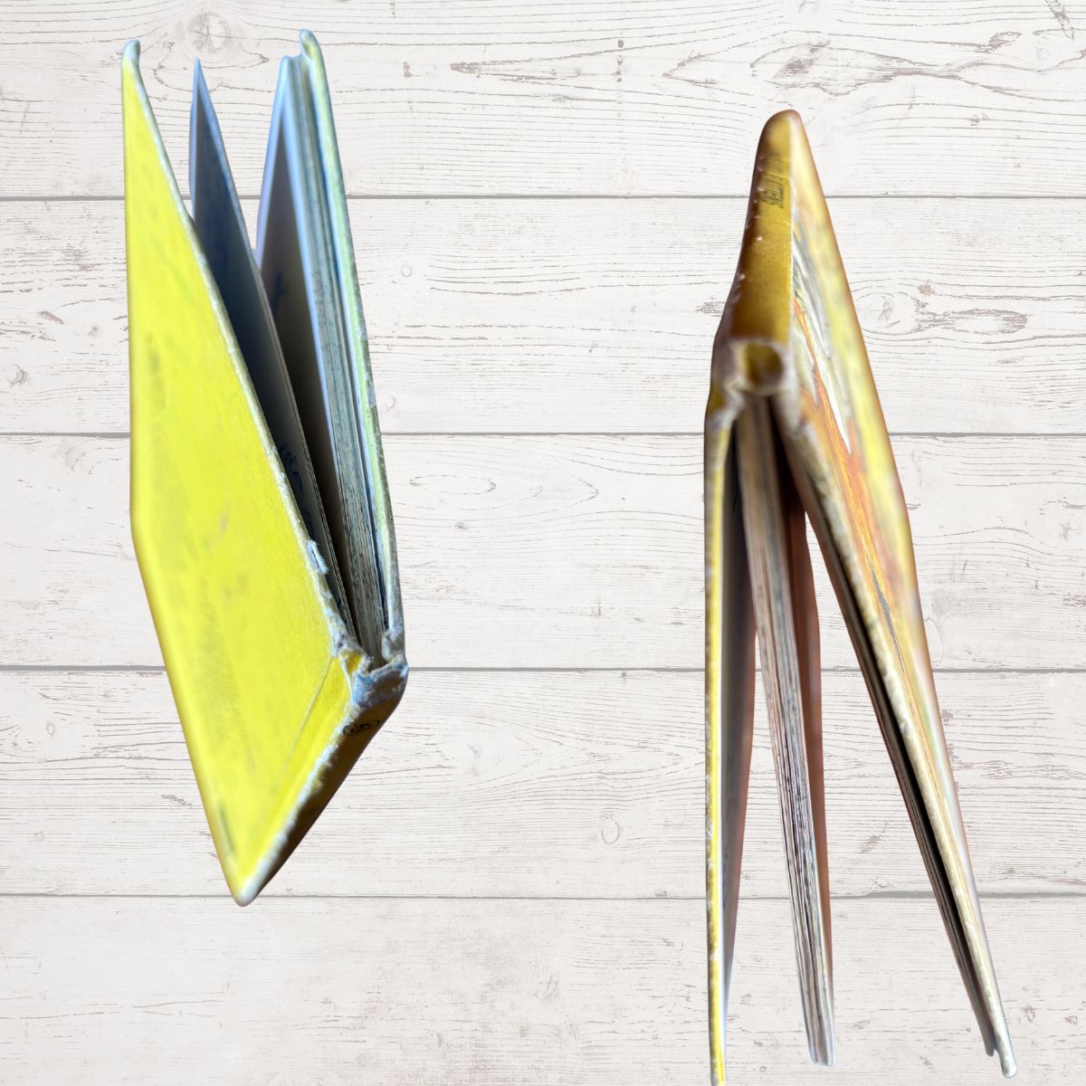 Two vintage yellow-bound books or notebooks standing open on a light wood plank background, one showing a bright yellow cover and the other showing a worn gold-toned cover.