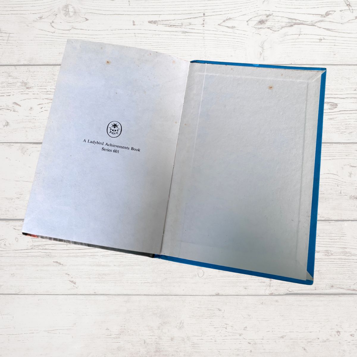 An open book with blank white pages on one side and a small printed title on the other, placed on a white wood surface. The book has a blue edge visible along the right side.