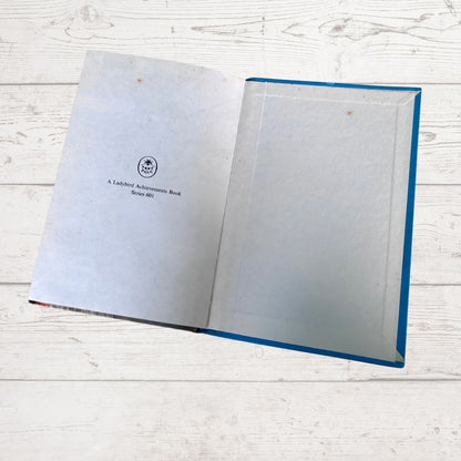 An open book with blank white pages on one side and a small printed title on the other, placed on a white wood surface. The book has a blue edge visible along the right side.