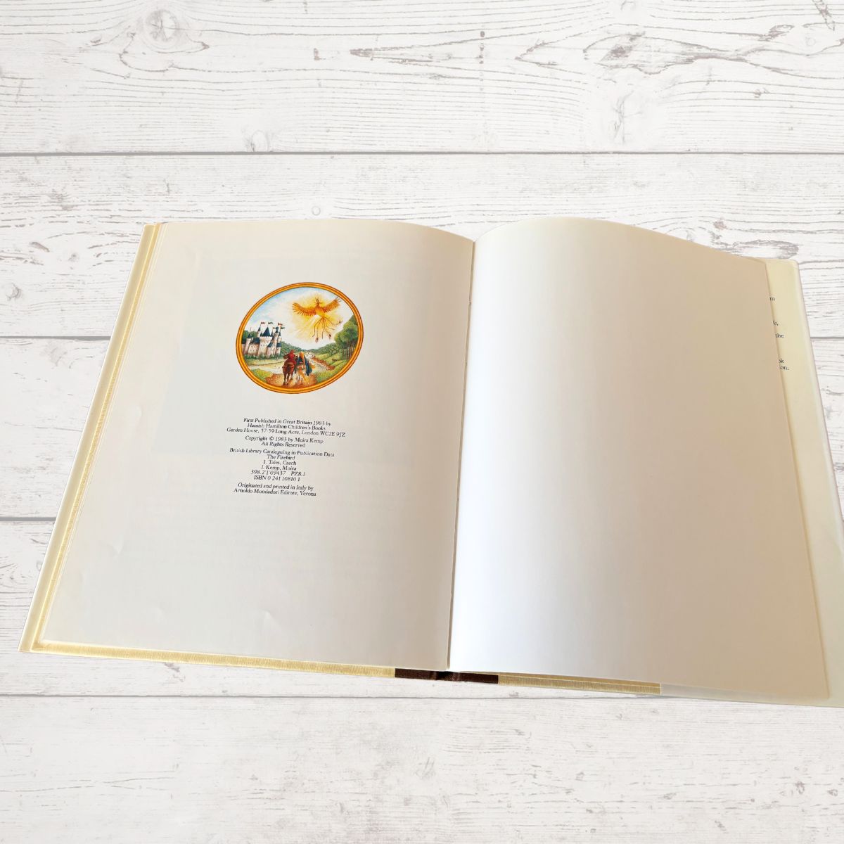 An open hardcover book laid flat on a white wood plank surface, showing cream-colored pages with a small circular vintage illustration and printed text on the left page.