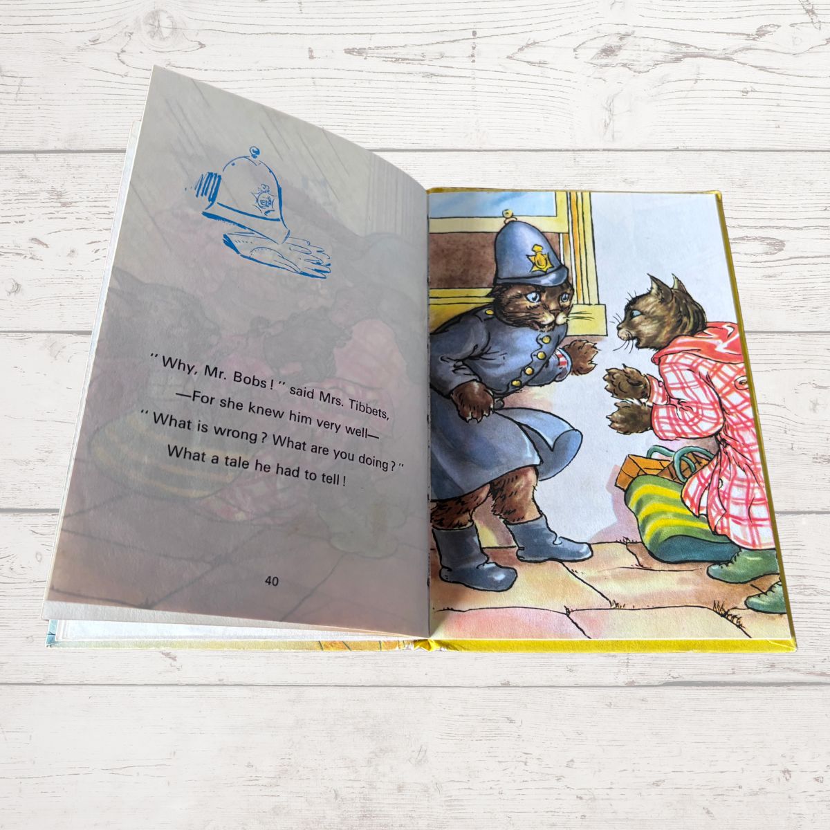 An open children's picture book lying on a white wood surface, showing a printed illustration of two cats dressed as a policeman and a victim, with text on the left page.