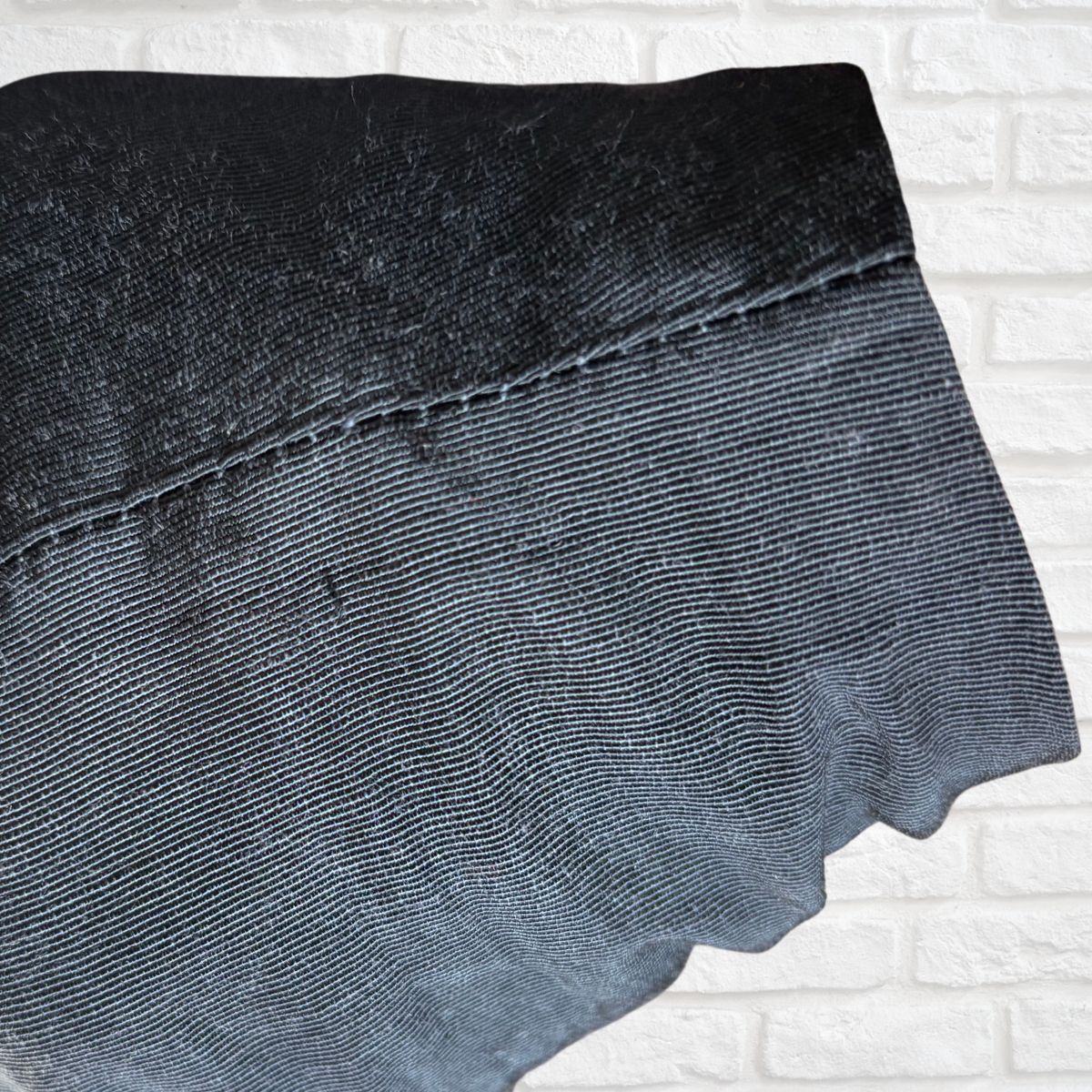 A close-up of a dark black textured fabric with a visible stitched seam, laid against a white brick wall background.