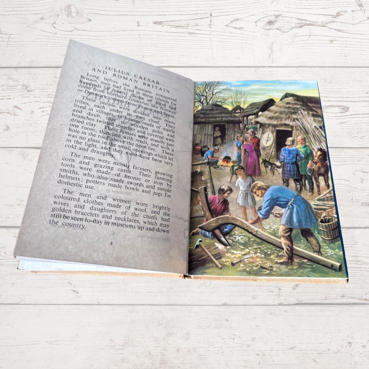 An open vintage book lying on a white wood surface, showing printed text on the left page and a colorful historical illustration on the right page depicting people in ancient clothing around thatched-roof huts.