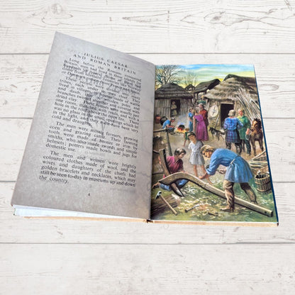 An open vintage book lying on a white wood surface, showing printed text on the left page and a colorful historical illustration on the right page depicting people in ancient clothing around thatched-roof huts.