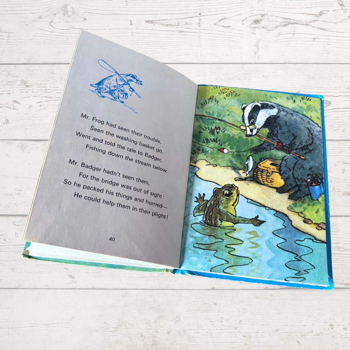 An open children's picture book lying on a light wood surface, showing a printed story page with text and a colorful illustration of animals by a river.