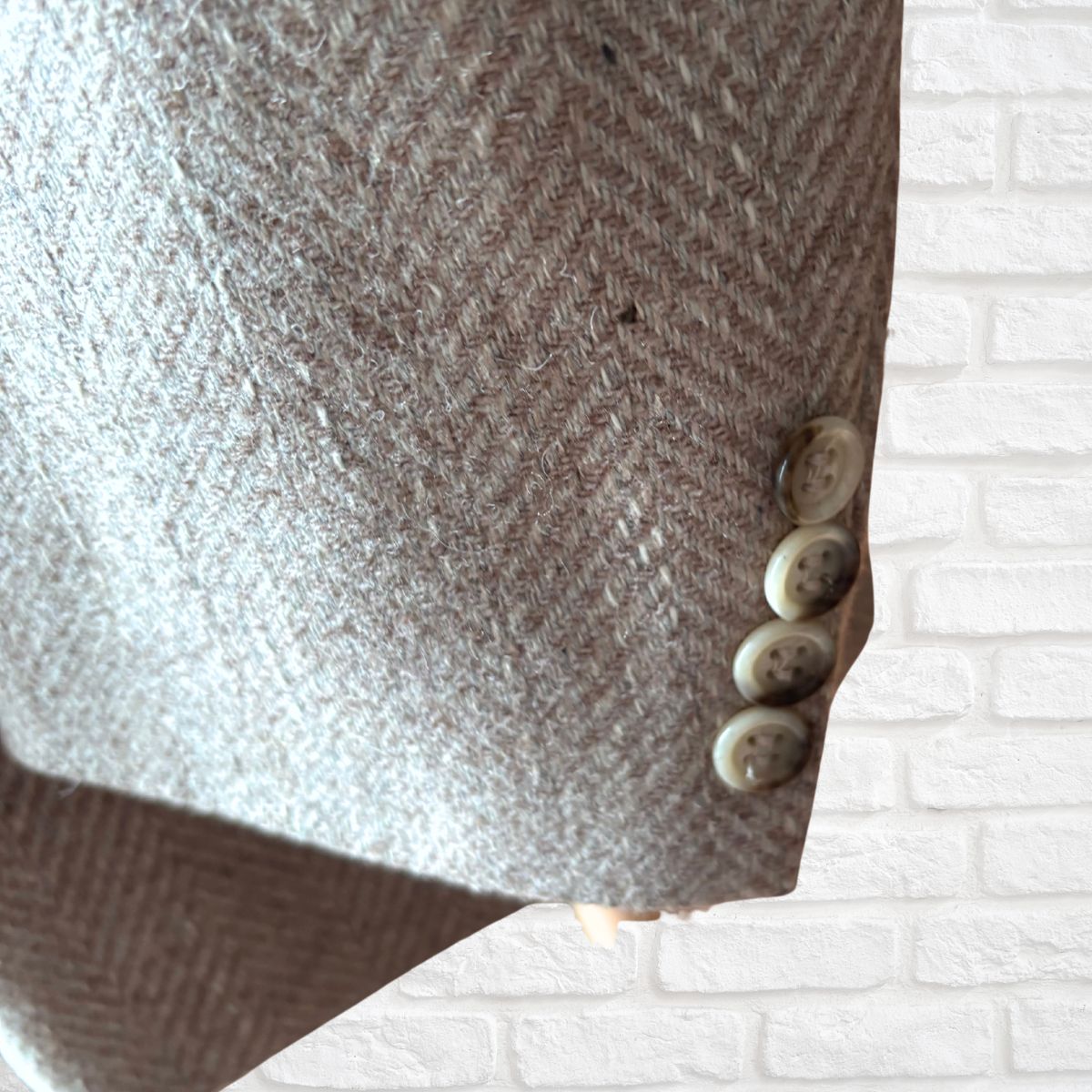 Close-up of a herringbone tweed jacket sleeve showing the textured fabric and four decorative buttons, set against a white brick wall background.