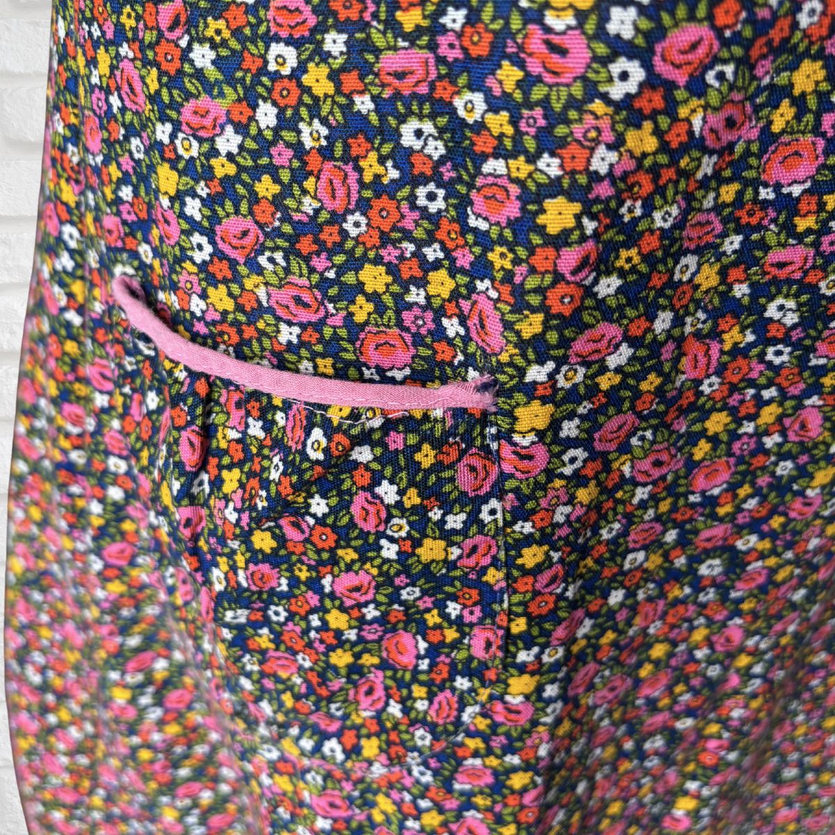 Close-up of a floral fabric with a small pink pocket and a pink trim detail, featuring a dense pattern of pink, yellow, white, and orange flowers on a dark navy background.