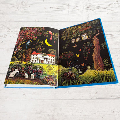 An open illustrated book with two illustrated spread pages, photographed on a white wood plank surface. The artwork depicts a dark woodland scene with a white house, birds, flowers, and a crescent moon; the book spine/border appears bright blue.