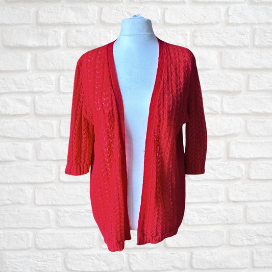 A red cable-knit open-front cardigan sweater with three-quarter sleeves, displayed on a light blue mannequin torso against a white brick wall background.