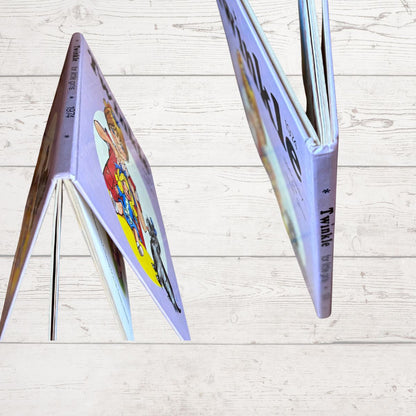 A children's book with a lavender-purple cover and colorful illustration, shown standing open on a light wood plank surface. The spine reads 'The Invisible Man' and the cover features a whimsical character design.