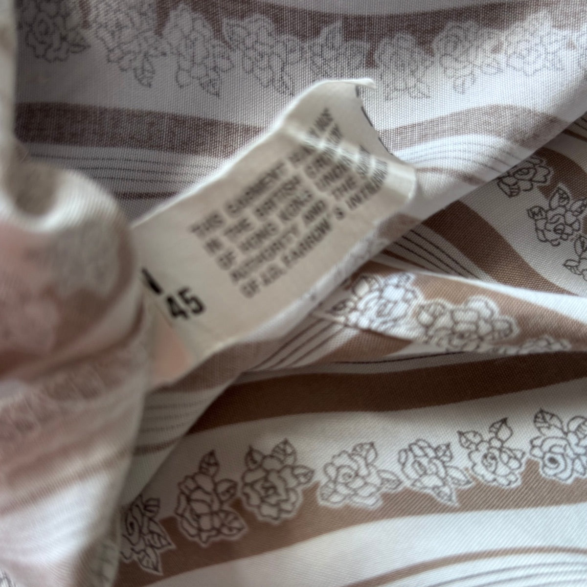 Close-up of a garment's inner label sewn onto patterned fabric. The fabric is cream and taupe with a floral line-art print.
