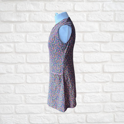 A sleeveless, mini length dress with a dense multicolour floral print, displayed on a light blue mannequin torso against a white brick wall background.