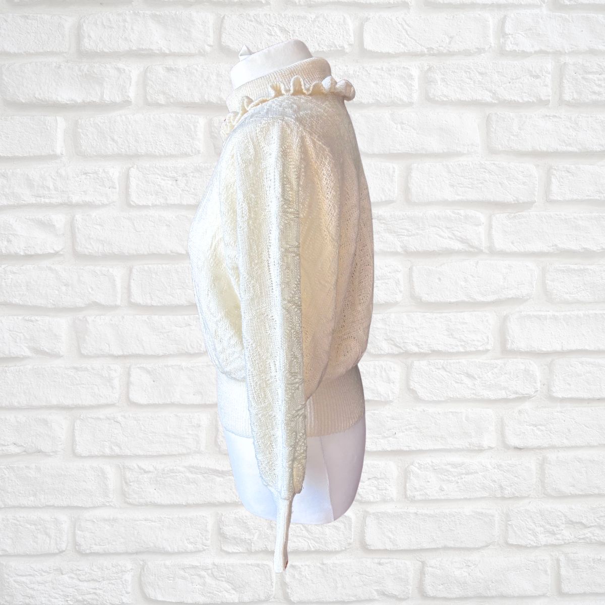 An ivory textured knit sweater with a ruffled mock neck, displayed on a white mannequin torso against a white brick wall background.