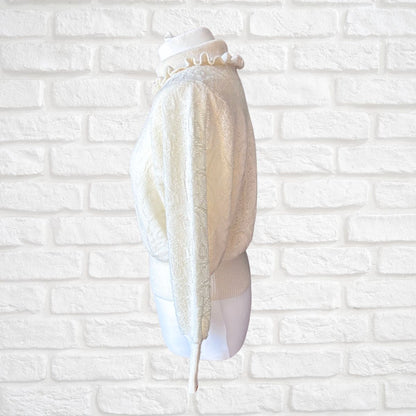 An ivory textured knit sweater with a ruffled mock neck, displayed on a white mannequin torso against a white brick wall background.