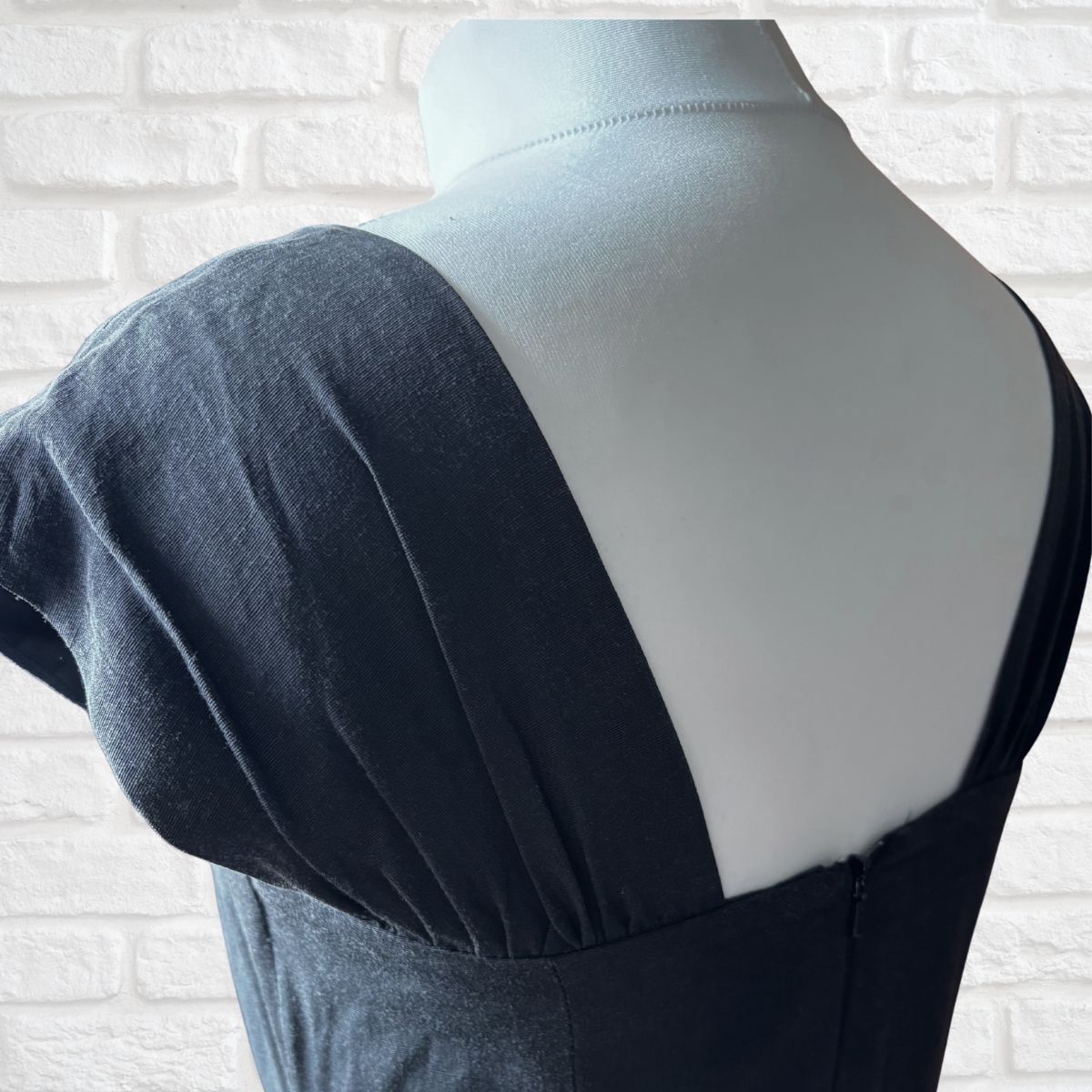 A close-up of a black dress displayed on a white mannequin torso, showing the back neckline and shoulder details against a white brick wall background.