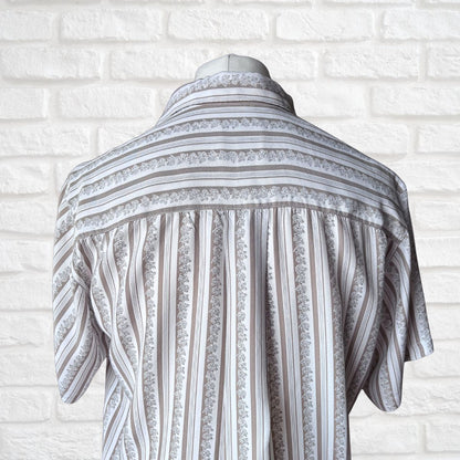 A short-sleeve button-up shirt with vertical stripes and floral lace-like bands, displayed on a mannequin torso against a white brick wall background.