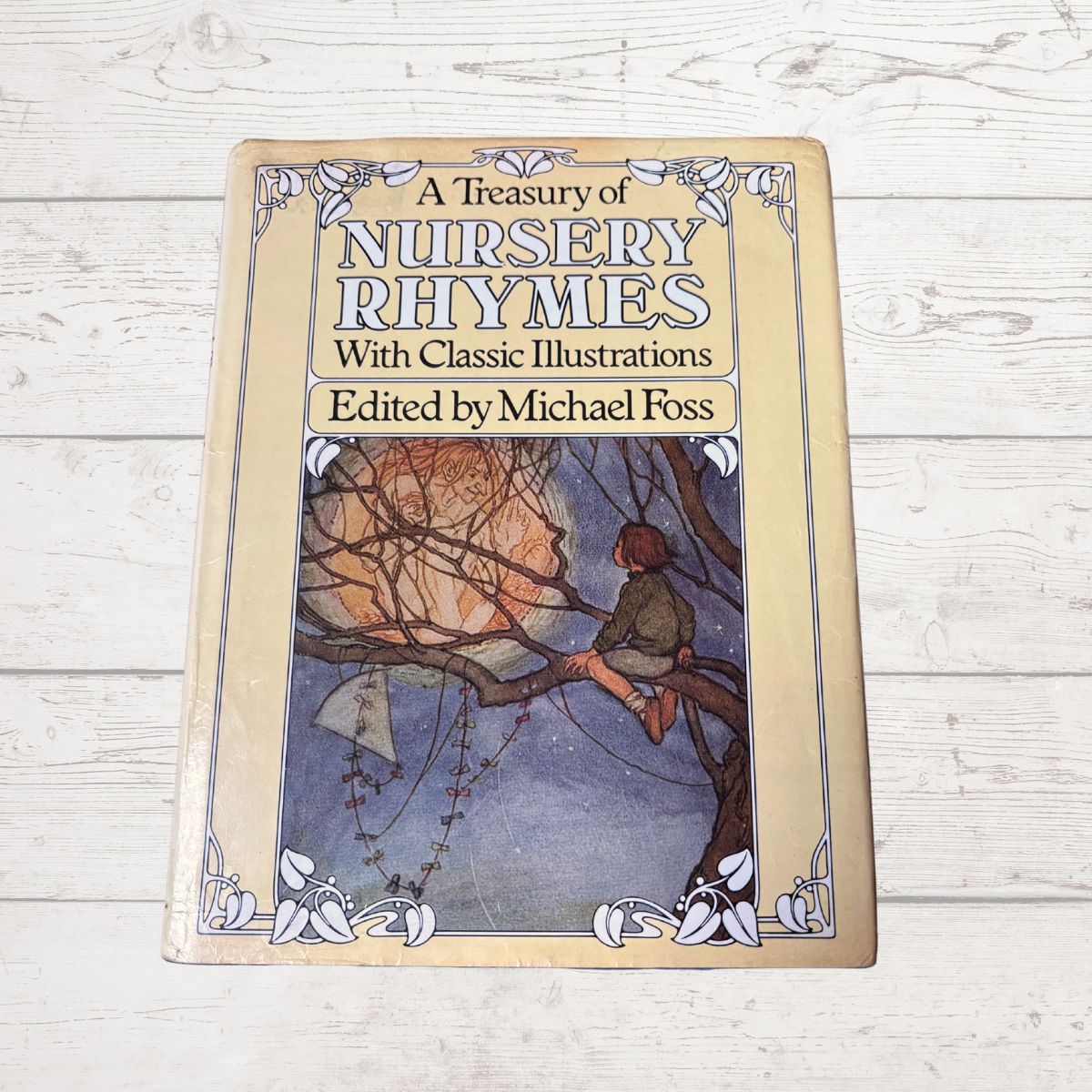 A hardcover book titled 'A Treasury of Nursery Rhymes With Classic Illustrations Edited by Michael Foss' placed on a white wood plank surface. The cover features a vintage-style illustration of a child sitting in a tree branch under a full moon.