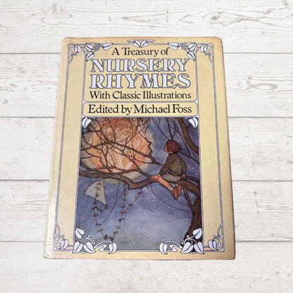 A hardcover book titled 'A Treasury of Nursery Rhymes With Classic Illustrations Edited by Michael Foss' placed on a white wood plank surface. The cover features a vintage-style illustration of a child sitting in a tree branch under a full moon.