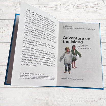 Open book showing a Ladybird Books reading scheme book titled 'Adventure on the Island' on a wooden surface.