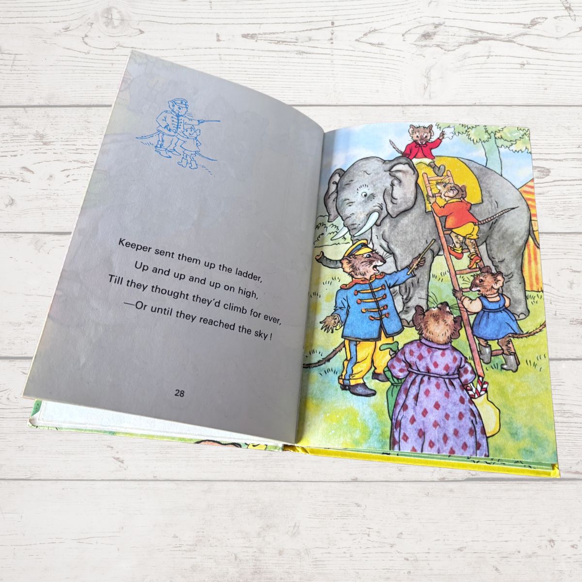An open children's book lying on a white wood surface, showing a two-page spread with text on the left page and a colorful illustration of children climbing an elephant on the right page.
