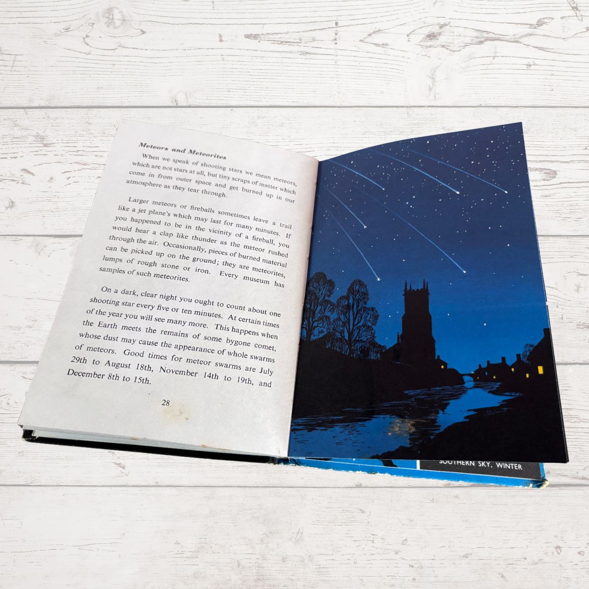 An open book lying on a white wood surface, showing printed text on the left page and a night sky photograph with a starry blue sky over a dark tree-lined scene on the right page.