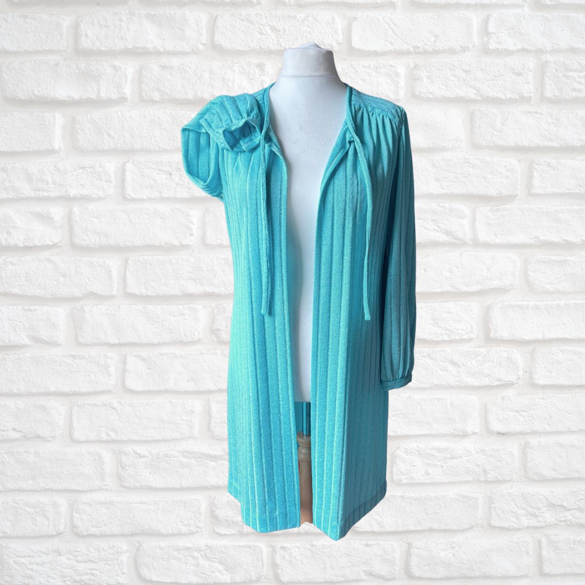 A turquoise blue open-front cardigan with ruffled short sleeves and a tie-neck detail, displayed on a white mannequin torso against a white brick wall background.