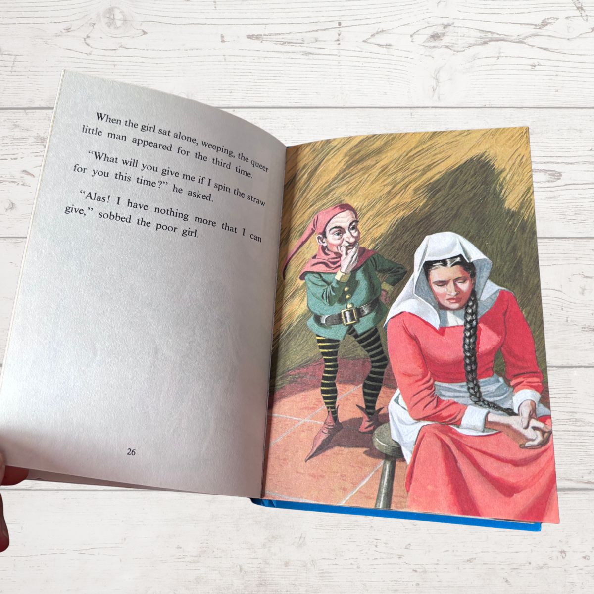 An open vintage children's book lying on a white wood surface, showing a printed story page with text on the left and a colored illustration on the right of a woman in a red dress and a man in a green coat.