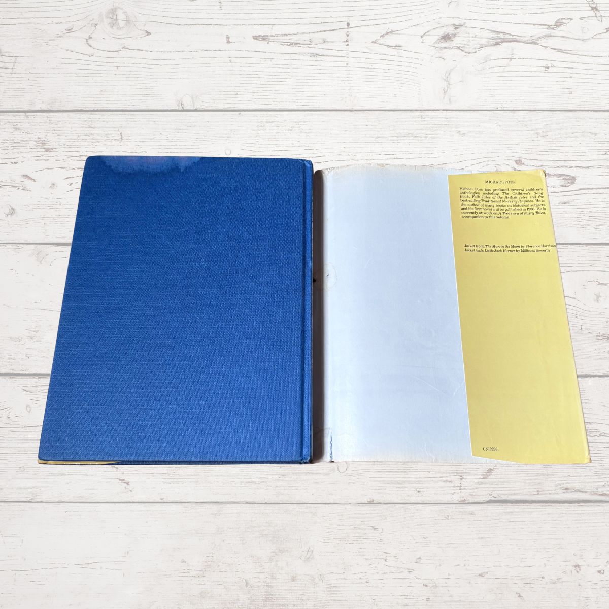 A vintage hardcover book or notebook lies open on a white wood plank surface. The left page is a solid blue cover, and the right page shows a yellow left margin with printed text and a white text block in the center.