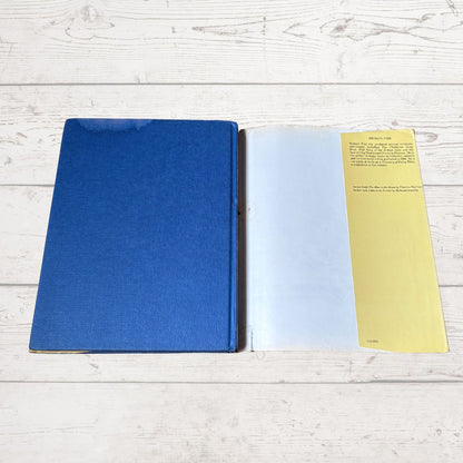 A vintage hardcover book or notebook lies open on a white wood plank surface. The left page is a solid blue cover, and the right page shows a yellow left margin with printed text and a white text block in the center.