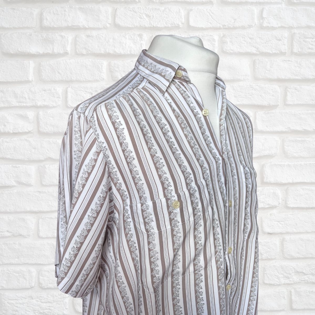 A short-sleeve button-up shirt with vertical stripes of brown, beige, and white, featuring a  floral pattern , displayed on a white mannequin torso against a white brick wall background.