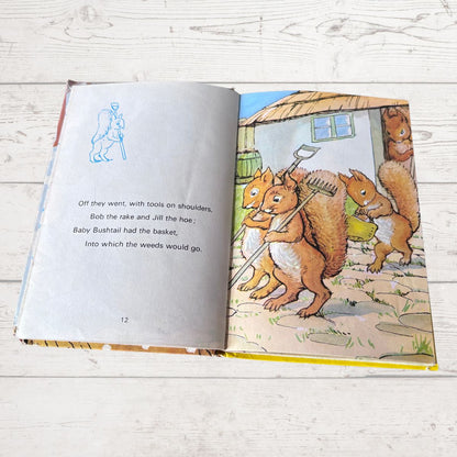 An open children's book lying on a white wood surface, showing a printed page with text and a colorful illustration of squirrels working in a garden.
