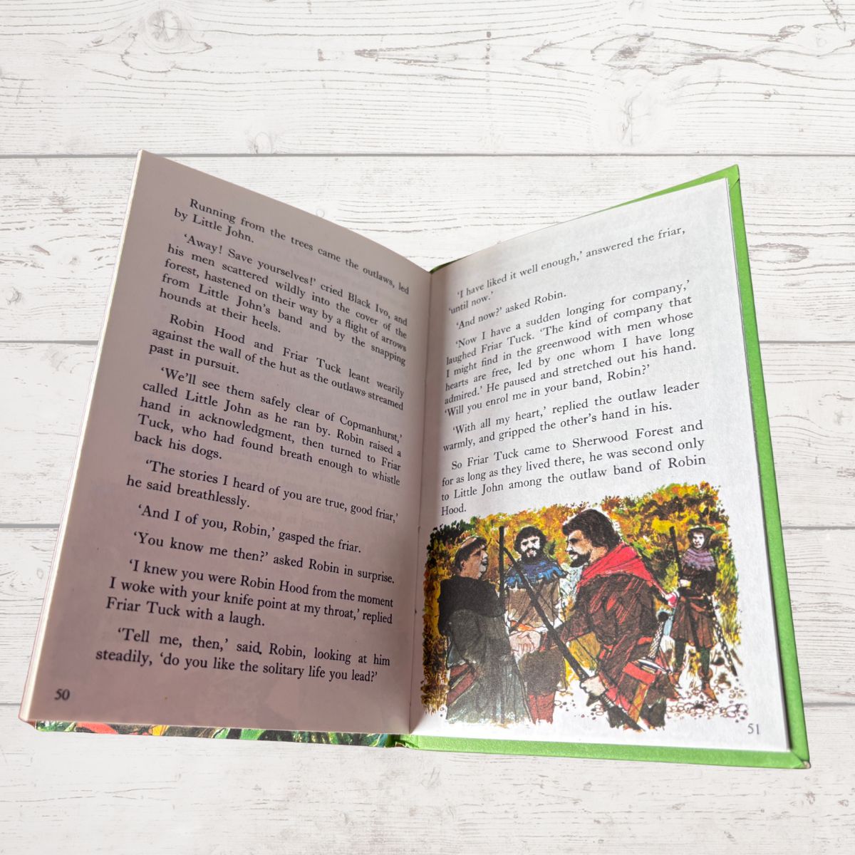 An open children's book lying on a white marble surface, showing printed text on the left page and a colorful illustration on the right page. The book has a green border and is photographed from above.