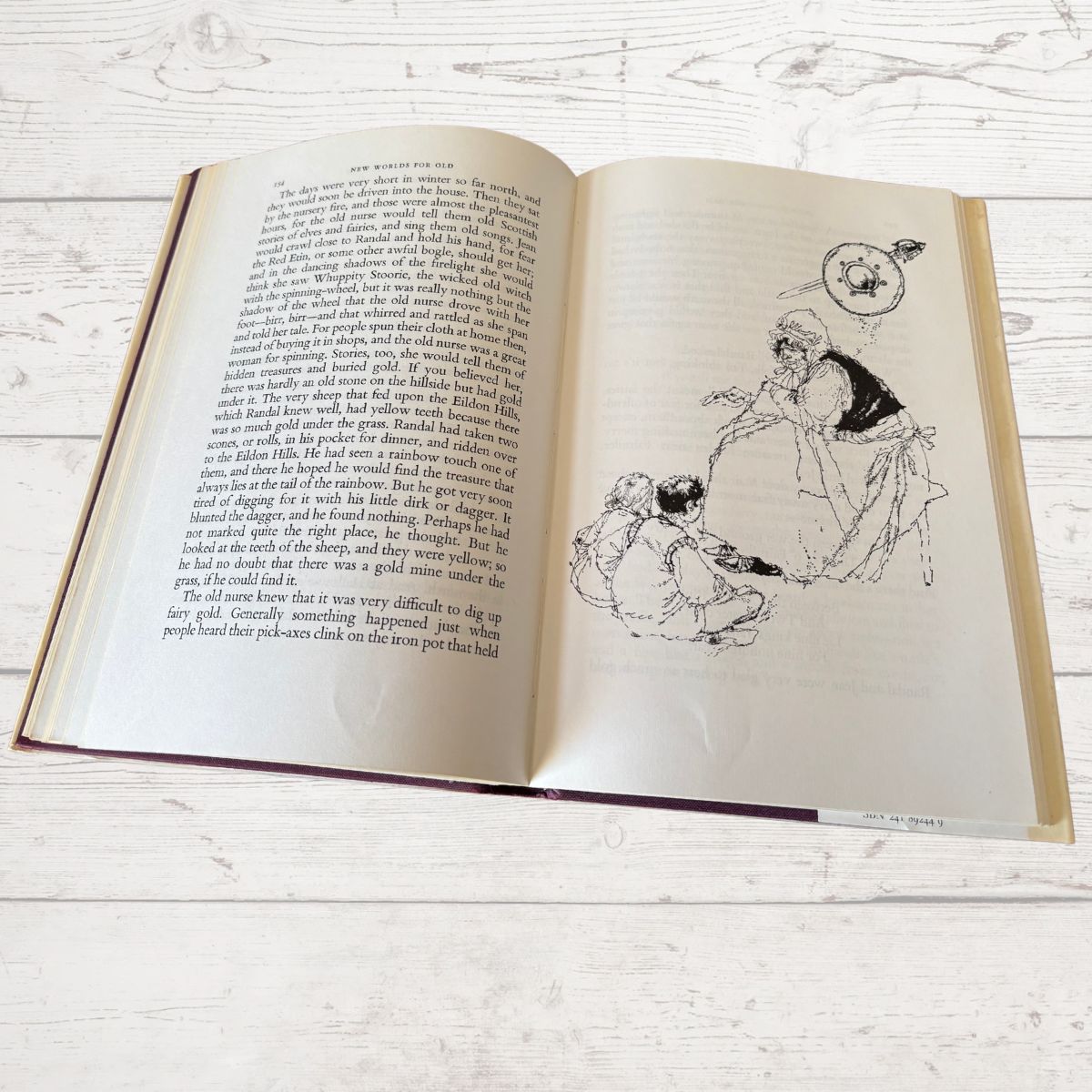 An open vintage book lying flat on a light whitewashed wooden surface, showing printed text on both pages and a black-and-white illustration on the right page.