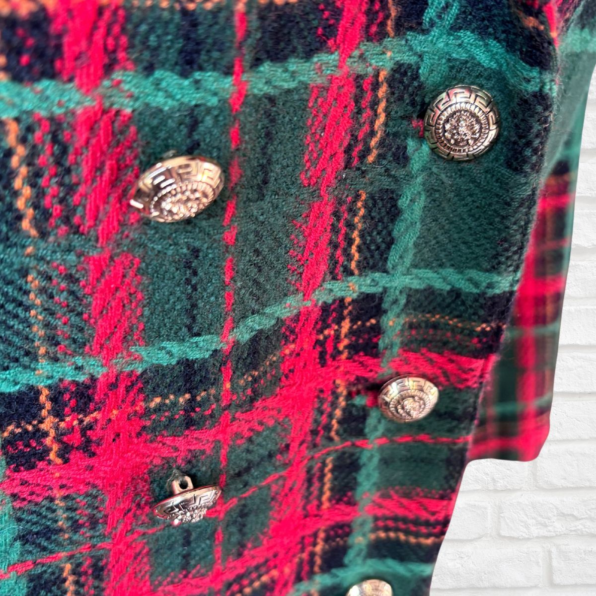 A close-up of a tartan plaid garment featuring a green, red, and black pattern with decorative silver-tone buttons, set against a white brick wall background.
