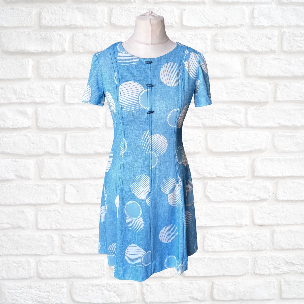 Blue dress with white circular patterns on a mannequin against a white brick wall.