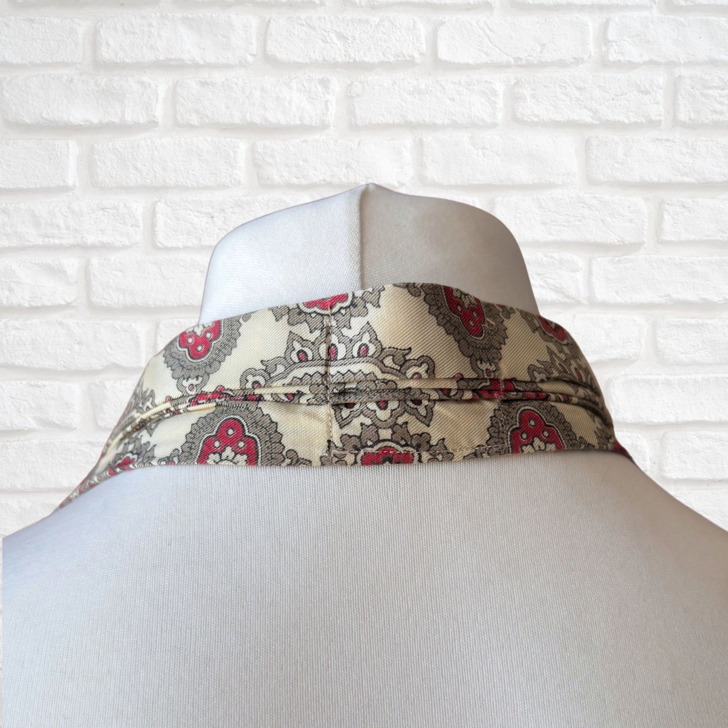 A vintage 60s mod style cream cravat with a paisley print featuring baroque style red, grey, and green patterns, displayed on a mannequin against a white brick wall background.