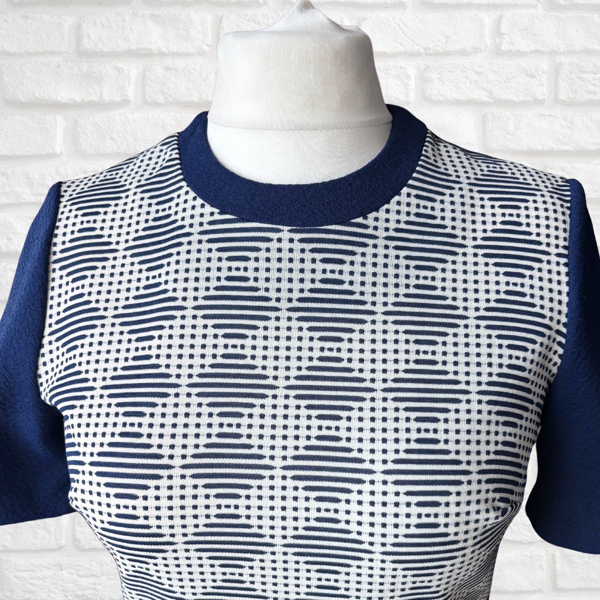 Mannequin wearing a blue and white patterned top against a white brick wall.