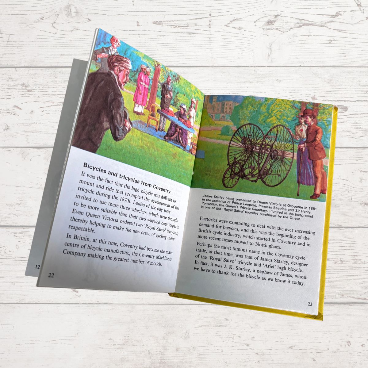 An open vintage-style book or booklet laid flat on a white wood surface, showing illustrated pages with text about bicycles and a colorful historical illustration of people riding bicycles.