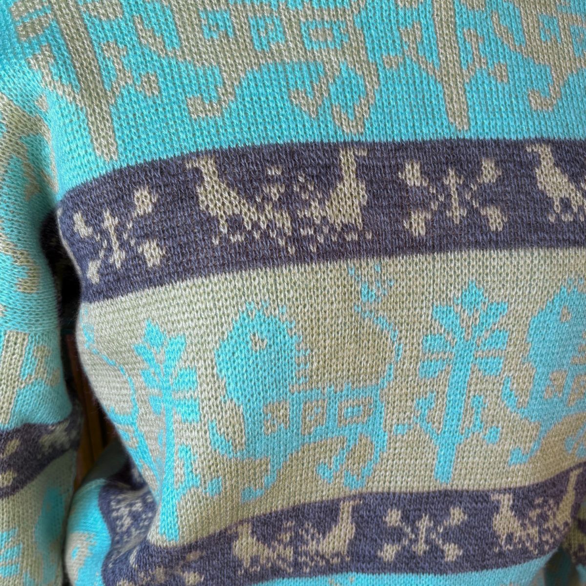 Close-up of a knitted sweater with horizontal bands of turquoise blue and warm beige, featuring a Nordic-style pattern with reindeer and snowflakes.