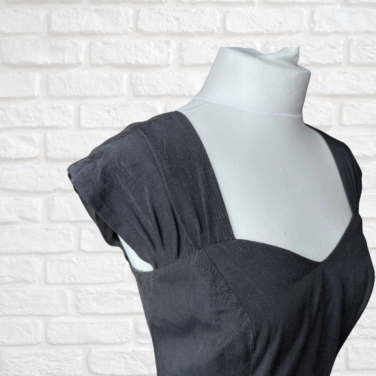 A black textured dress displayed on a white mannequin torso against a white brick wall background.