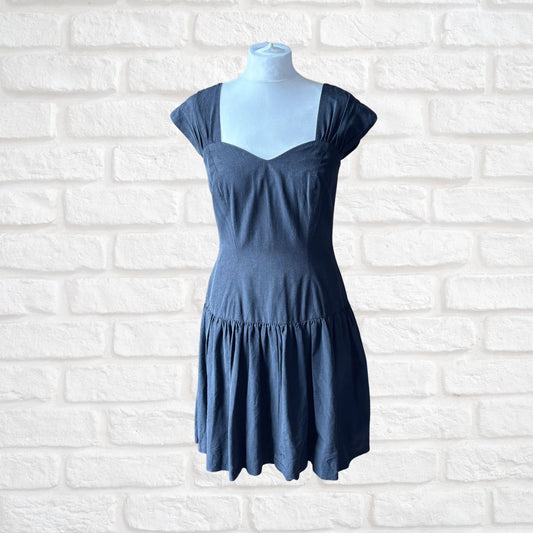 A black tiered mini dress with cap sleeves and a scoop neckline, displayed on a light blue mannequin torso against a white brick wall background.