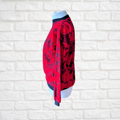 A red long-sleeve blouse with a black floral pattern, displayed on a light blue mannequin torso against a white brick wall background.