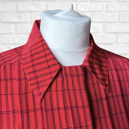 A red collared shirt dress with a black windowpane plaid pattern, displayed on a white fabric mannequin torso against a white brick wall background.