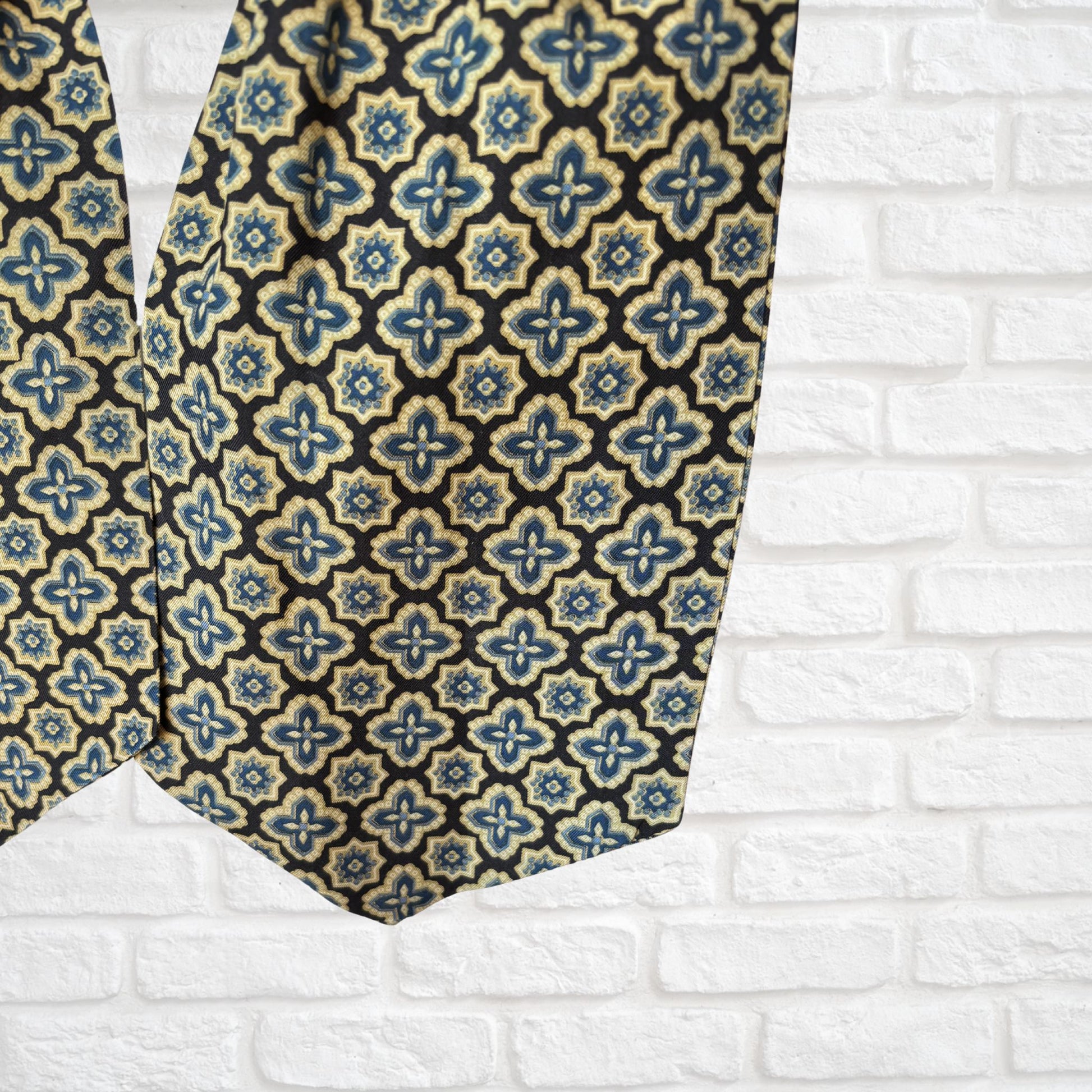 Close up of a vintage Austin Reed cravat featuring a black necktie with a cream, gold, and blue abstract  floral print, displayed on a mannequin against a white brick wall background.