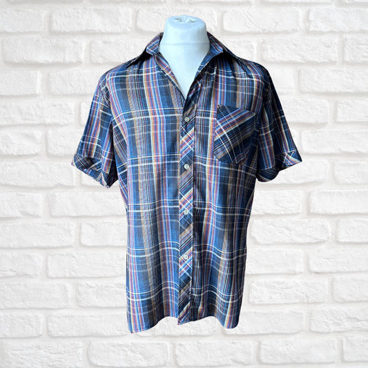 A short-sleeve plaid button-up shirt in blue, navy, red, and white, displayed on a light blue mannequin torso against a white brick wall background.