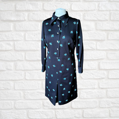 Black dress with turquoise patterns on a mannequin against a white brick wall.