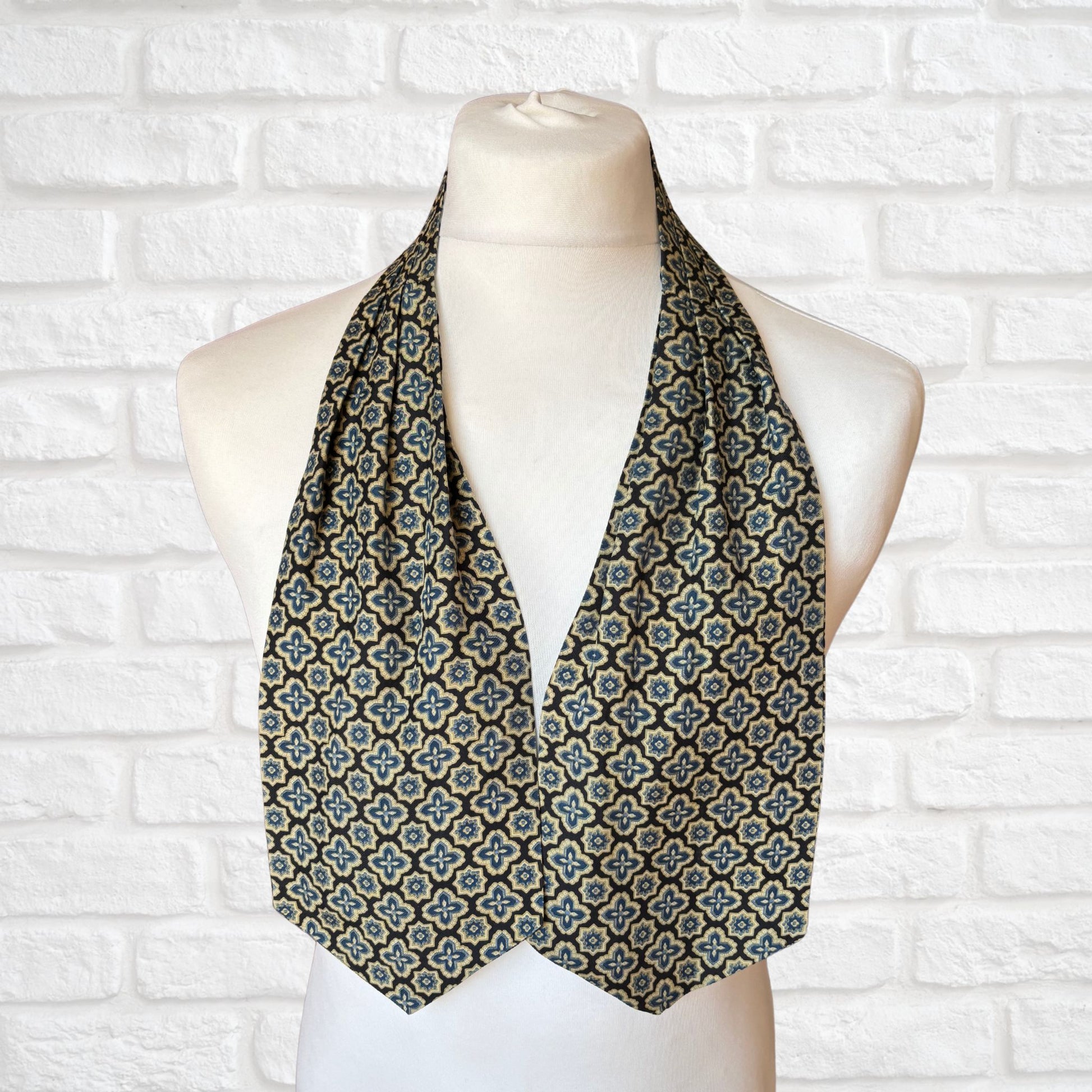 A vintage Austin Reed cravat featuring a black necktie with a cream, gold, and blue stylized floral print, displayed on a mannequin against a white brick wall background.