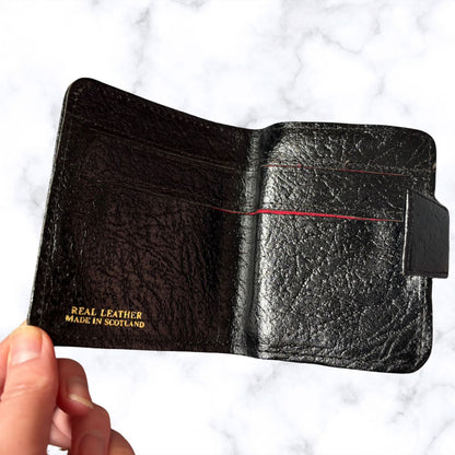 Black leather wallet held by a hand with 'Real Leather Made in Scotland' text on a white marble background.