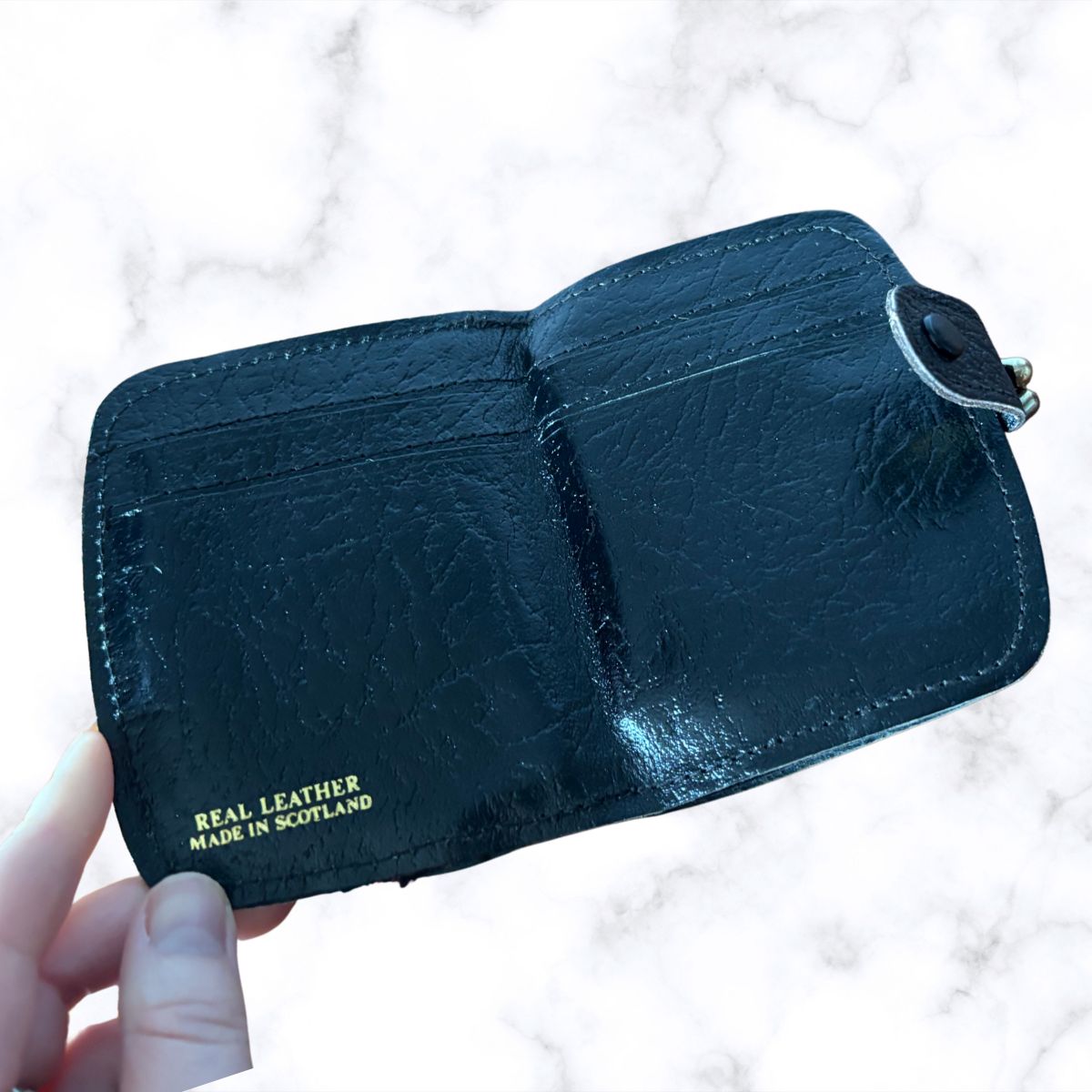 Black leather wallet with 'Real Leather Made in Scotland' text held by a hand on a light background