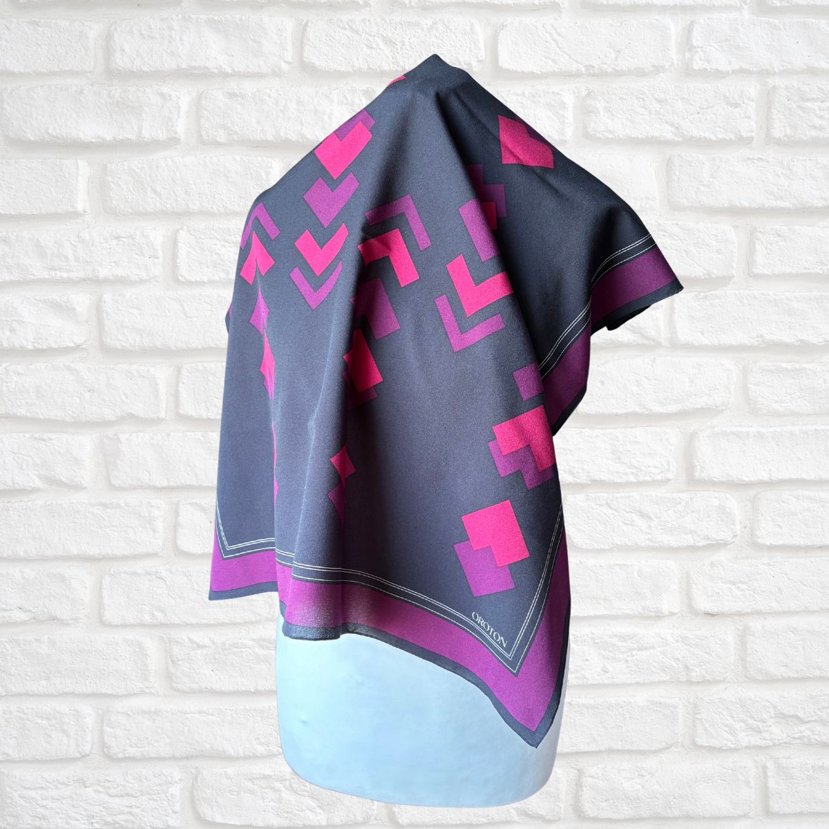 Scarf with pink and purple geometric pattern draped over a light blue surface against a white brick wall.